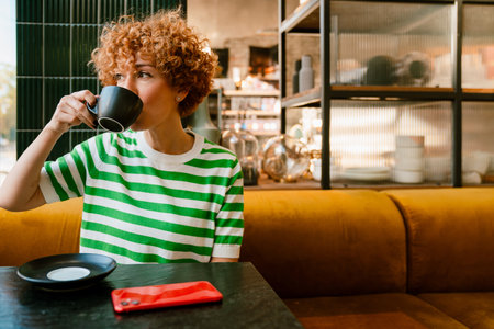Mid white woman with curly hair drinking coffee while sitting by table in cafeの写真素材