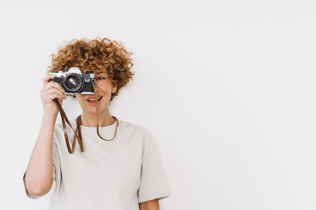 Ginger curly woman wearing t-shirt taking photo on retro camera isolated over white backgroundの写真素材