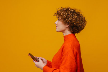 Profile of ginger mature woman using smartphone while standing isolated over yellow wallの写真素材