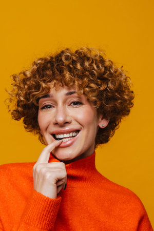 Mature ginger woman touching her lips and looking at camera while standing over isolated yellow...