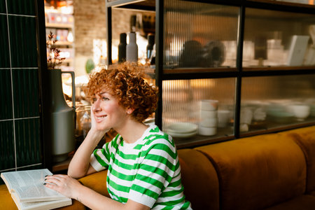 Mid white woman with curly hair reading book while sitting by table in cafeの写真素材