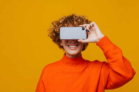 Cheerful mature woman covering her face with mobile phone isolated over yellow studio background