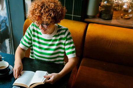 Young ginger curly woman in eyeglasses reading book while drinking coffee in cafeの写真素材
