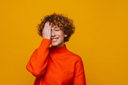Cheerful middle-aged woman covering her face while standing isolated over yellow studio backkground