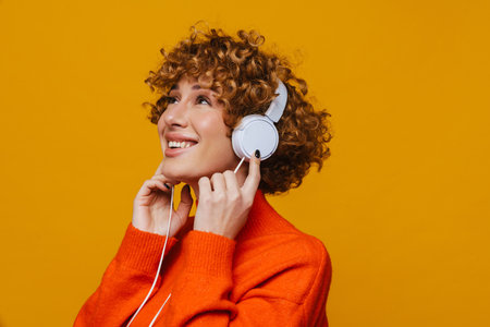 Middle-aged ginger woman listening music with headphones looking up isolated over yellow backgroundの写真素材