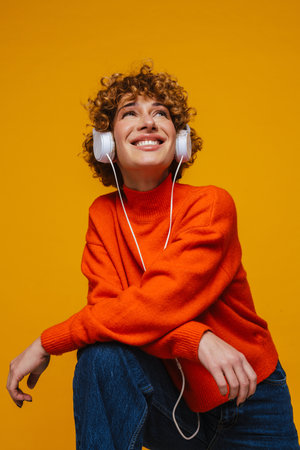 Cheerful middle-aged ginger woman listening music with headphones and looking up isolated over yellow backgroundの写真素材