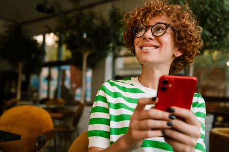 Mid white woman with curly hair smiling and using cellphone while sitting in cafeの写真素材
