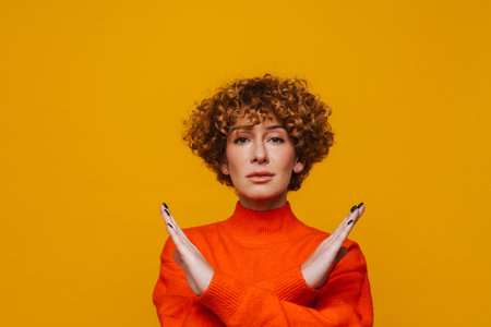 Middle-aged woman showing crossed hands gesture and looking at camera while standing isolated over yellow backgroundの写真素材