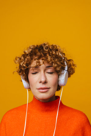 Ginger woman in headphones listening music while posing on camera isolated over yellow background