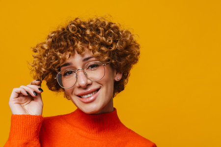 Joyful middle-aged ginger woman in glasses touching hair isolated over yellow studio backgroundの写真素材