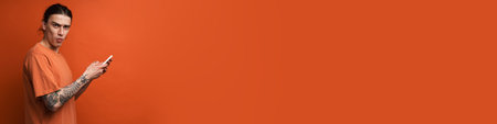 Young white man expressing surprise while using cellphone isolated over orange background