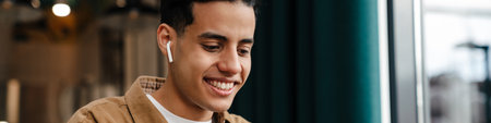 Young smiling hispanic man student sitting at the cafe table with laptop computer indoors, holding mobile phone, watching videoの写真素材