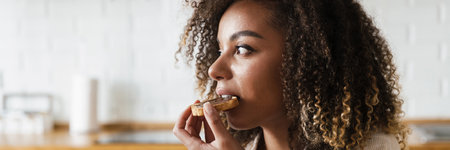 The side view of a woman eating a toast with a chocolate paste while sitting in the white kitchenの写真素材
