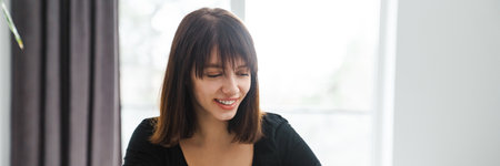 Happy young white woman looking at mobile phone while sitting at the kitchen table at home