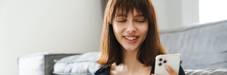 Smiling young woman shopping online via mobile phone holding credit card while sitting at the couch at homeの写真素材