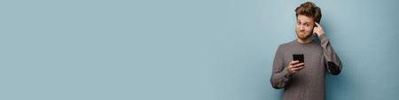 Portrait of a confused pensive young white casual man standing over gray background holding mobile...