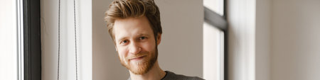 Ginger young man using mobile phone while leaning on wall indoors
