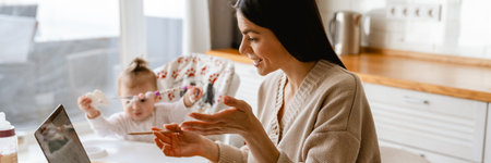 Young mother working with laptop while keeping her baby in kitchen at home