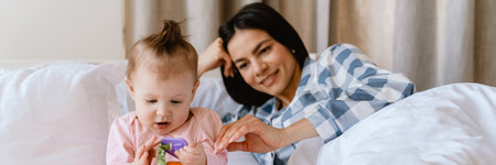 Young mother smiling and playing with her baby while lying in bad at home