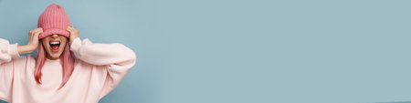 Young white woman with pink hair screaming and pulling hat down over her face isolated over blue...