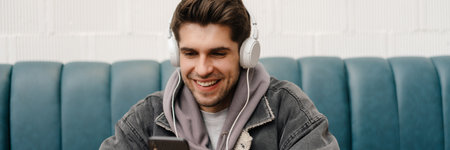Smiling young casual man in headphones listening to music, holding mobile phone while sitting in...