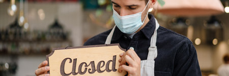 Young man waiter in apron and face mask showing closed sign while standing in cafe