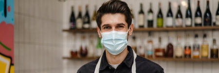 Young white brunette man in apron and protective mask holding cup of coffee, thumbs up