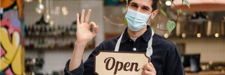 Young man waiter in apron showing open sign while standing indoors the cafe, ok gesture