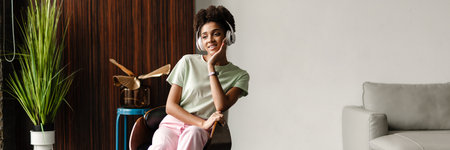 Young black woman listening music with headphones while sitting in armchair at homeの写真素材