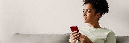 Young black woman using mobile phone while sitting on couch at homeの写真素材