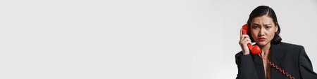 Brunette asian woman wearing jacket frowning while talking on retro phone isolated over white...
