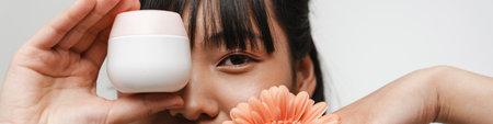 Young asian woman posing with face cream and gerbera flower isolated over white backgroundの写真素材