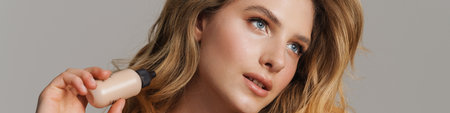 Half-naked blonde woman looking aside while showing cosmetic product isolated over grey background