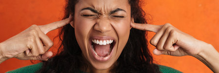 Frowning young african woman in casual wear standing over orange background, screaming, blocking...