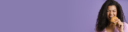 Young black woman with curly hair laughing while eating hamburger isolated over purple background