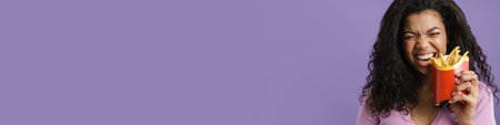 Young black woman smiling while eating french fries isolated over purple backgroundの写真素材