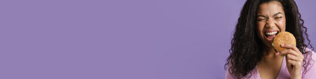Young black woman with curly hair laughing while eating hamburger isolated over purple backgroundの写真素材
