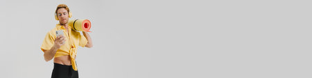 Young white gay man carrying fitness mat listening to music with headphones standing over gray wall...