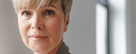 European mature woman looking at camera while standing by window indoors