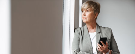 European mature woman using mobile phone while leaning on wall indoorsの写真素材