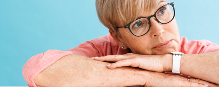 European mature woman looking aside while leaning on table isolated over blue background