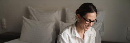 Smiling mid aged brunette white businesswoman sitting on bed in a hotel room holding mobile phone