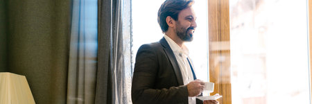 Smiling mid aged white man in formal wear standing at the window in the hotel room holding cupの写真素材