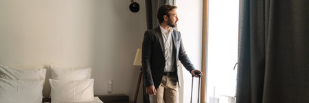 Mid aged white man in formal wear standing at the window in the hotel room holding suitcase