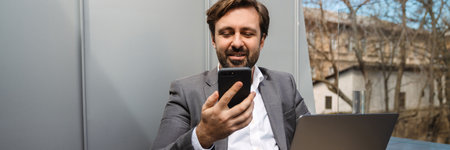 Smiling mid aged bearded businessman sitting on a balcony working on laptop computer