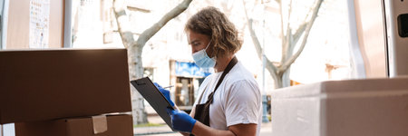 Young white cafe worker in face mask writing while taking delivery outdoors