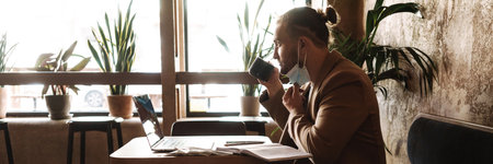 Young white man in face mask using laptop while drinking coffee in cafeの写真素材