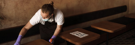 A male waiter in a protective disposable mask and uniform wiping a table in a cafe