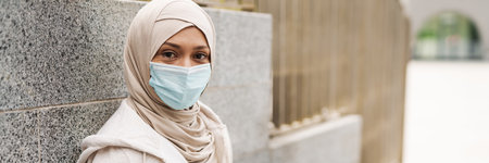 Mid aged muslim woman in protective medical mask holding mobile phone leaning on a wall on a streetの写真素材