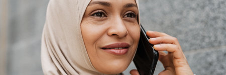 Middle eastern woman in hijab smiling while talking on cellphone outdoorsの写真素材